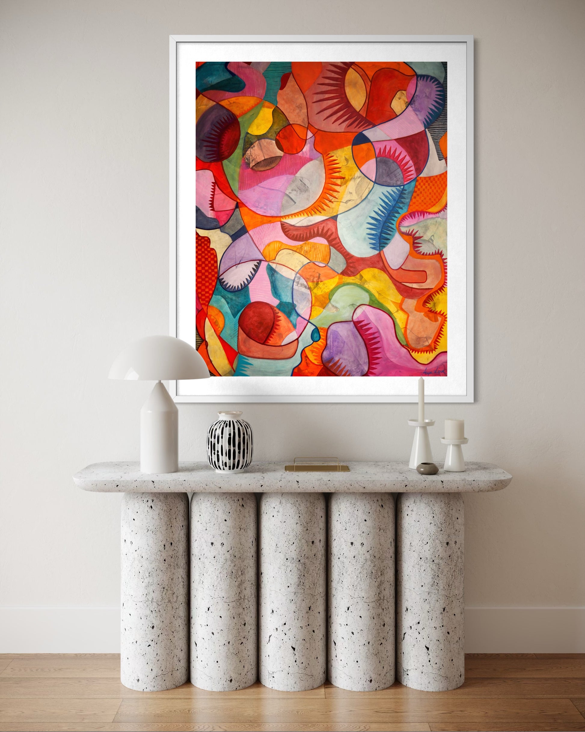 A framed abstract art print with a colorful, organic pattern in shades of red, orange, pink, yellow, green, and blue, displayed on a beige wall above a modern white console table with cylindrical legs.